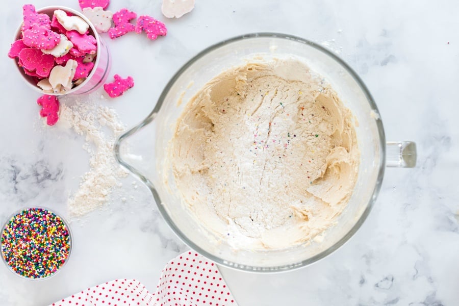 Flour and sprinkles being added to cookie batter in mixing bowl, with a small bowl of cookies, and sprinkles on the side