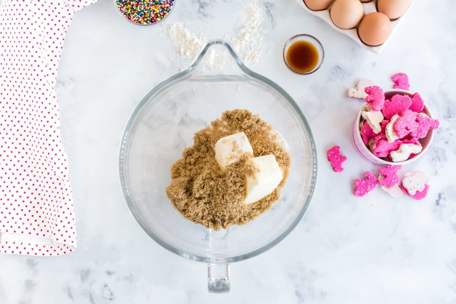 Sugars and butter in mixing bowl with a side bowls of animal cookies, vanilla, sprinkles and eggs in carton