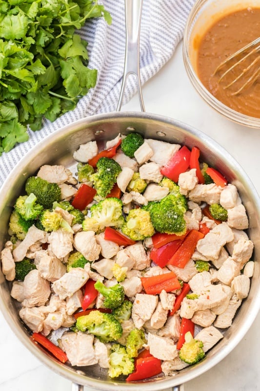 cut up chicken and vegetables in a cooking pan, in the back, a bowl of peanut sauce and a bunch of cilantro