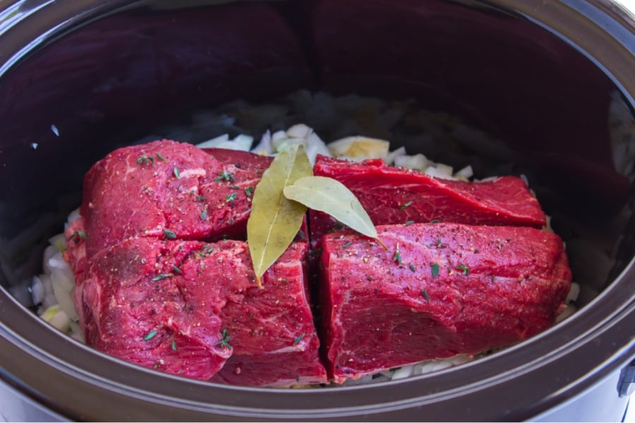 chopped veggies, quartered beef topped with bay leaves in a black slow cooker