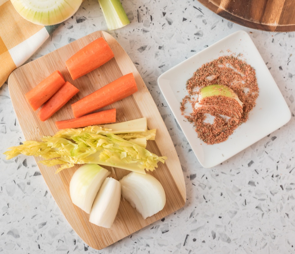 onions, carrots and celery on a cutting board, with a side of seasoning with a cut onion in it