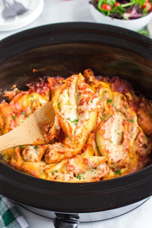 Crockpot Stuffed Shells in a slow cooker with a wooden spoon scooping up a serving
