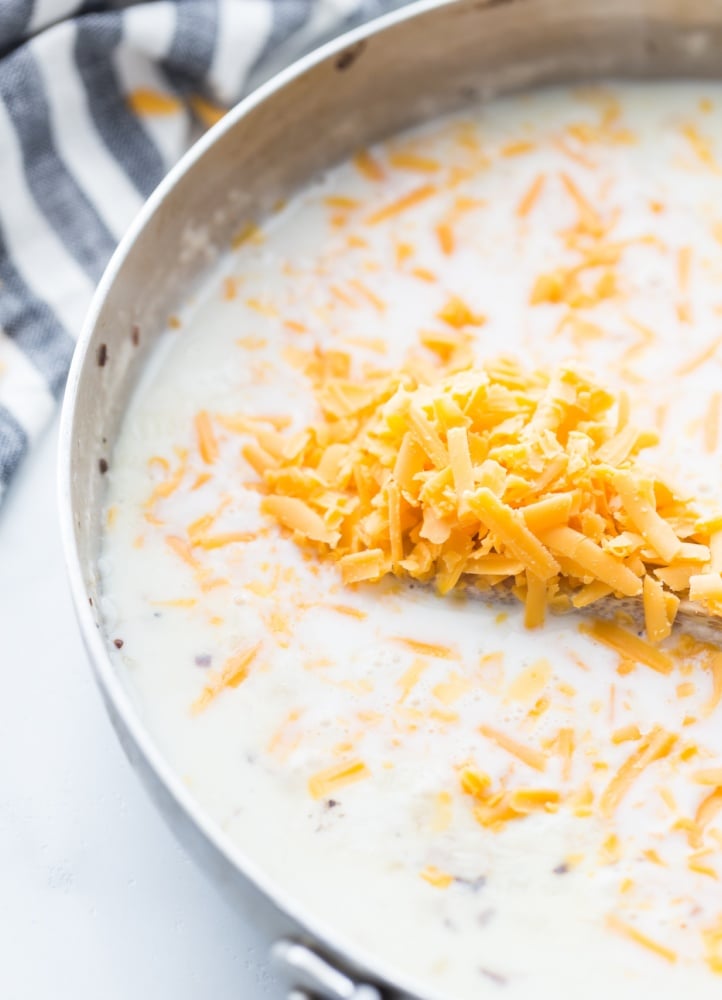 creamy sauce in a pot with shredded cheese being added in
