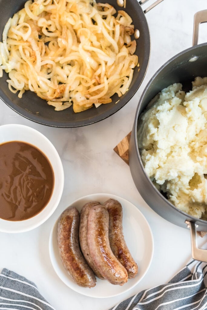 sliced onions cooking in pan, cooked sausage on a plate and a pot of mashed potatoes