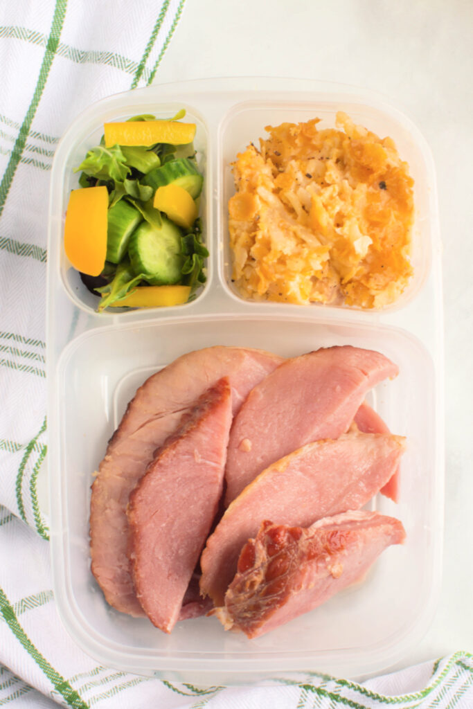  Classic Cheesy Funeral Potatoes in a lunchbox with ham slices and vegetables