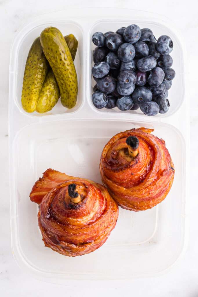 two bacon wrapped chicken drumsticks in a lunchbox with pickles and blueberries