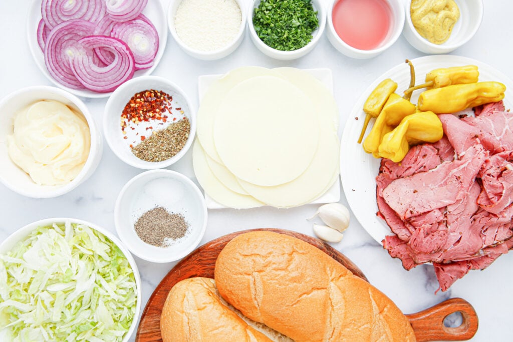 Grinder sandwich ingredients including hoagie rolls, pastrami, provolone cheese, shredded lettuce, pepperoncini, and red onion